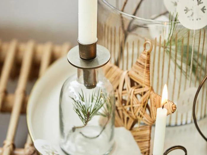 Et hvidt lys i en glasholder med en fyrrekvist, en flettet stjernepynt og et andet tændt lys på en træbakke.