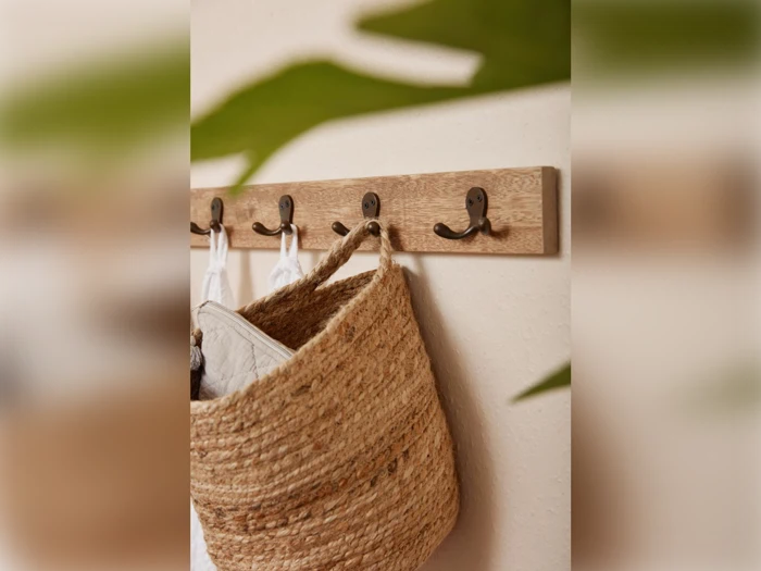 En vævet stråtaske og hvide klude hænger på et vægstativ af træ med metalkroge; grønne blade er slørede i forgrunden.
