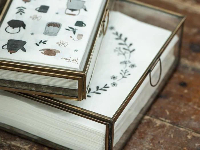 To glasæsker med messingkanter, som hver indeholder en stak hvidt papir med fine sorte blomster- og kaffeillustrationer, hviler på en træplade.