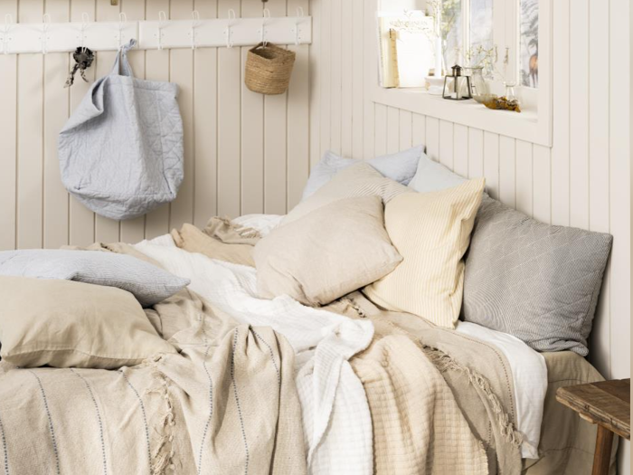 En hyggelig seng med beige, cremefarvede og grå puder og tæpper ved siden af et vindue, hvor sollyset strømmer ind. Lyse trævægge og enkel indretning skaber en varm og indbydende atmosfære.