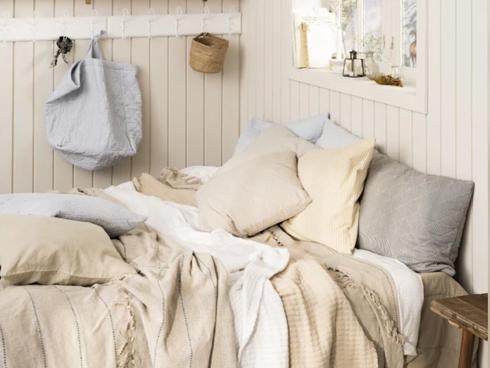 En hyggelig seng med beige, cremefarvede og grå puder og tæpper ved siden af et vindue, hvor sollyset strømmer ind. Lyse trævægge og enkel indretning skaber en varm og indbydende atmosfære.