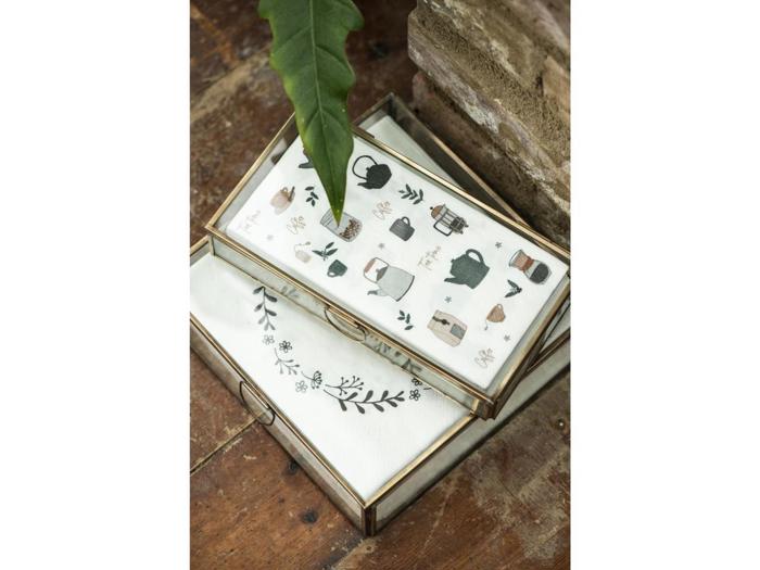 To glasmontrer med guldkanter, som indeholder hvide klude dekoreret med botaniske illustrationer og køkkenmotiver, står på en rustik træplade med et stort grønt blad i nærheden.