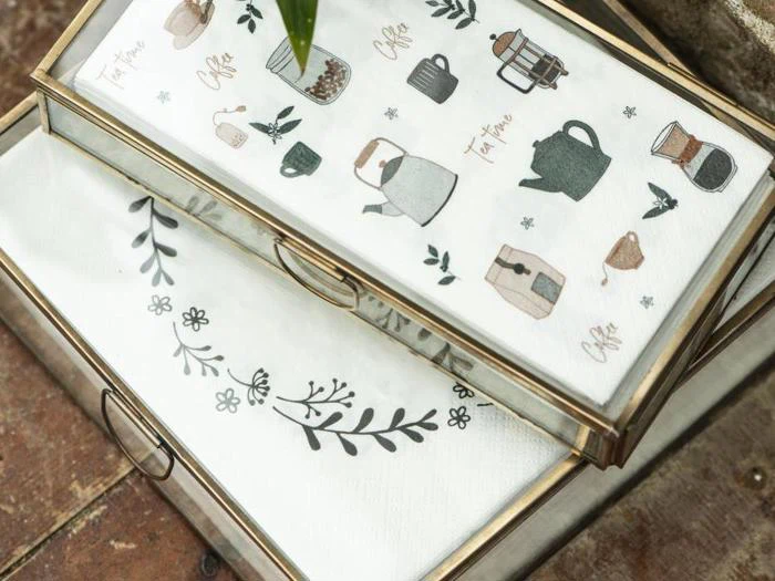 To glasbakker rummer servietter; den ene serviet viser illustrationer af kaffe og te, den anden har et enkelt sort blomsterdesign.
