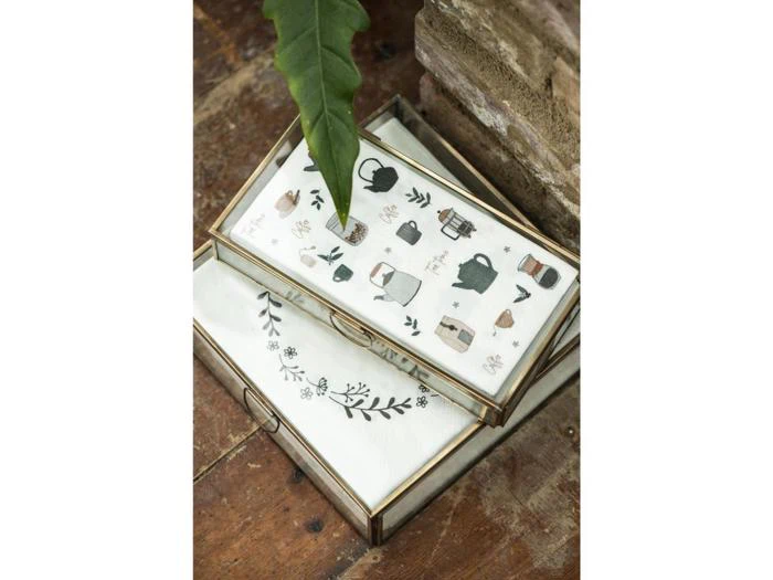 To glasmontrer med guldkanter, som indeholder hvide klude dekoreret med botaniske illustrationer og køkkenmotiver, står på en rustik træplade med et stort grønt blad i nærheden.