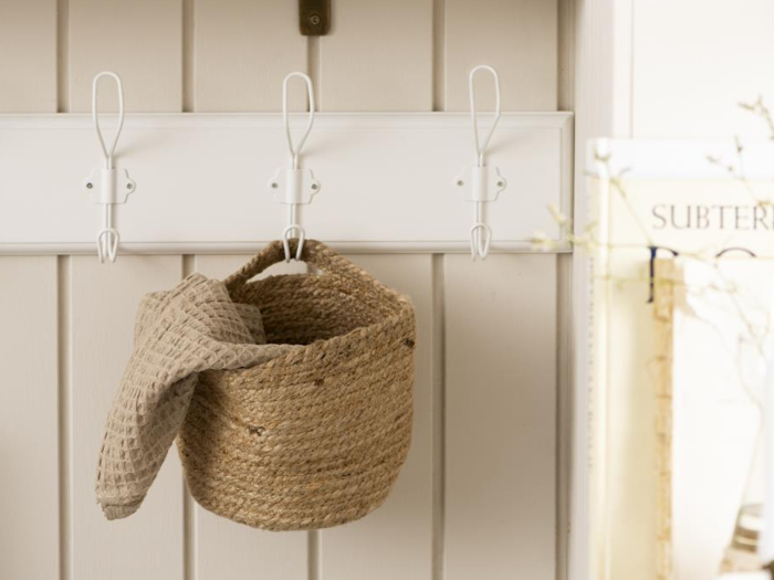En flettet kurv med et struktureret beige stof indeni hænger på en hvid knagerække, der er monteret på en lys trævæg.