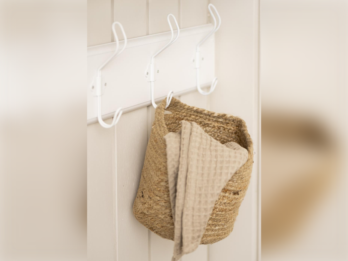 En flettet kurv og et beige tekstureret håndklæde hænger på hvide kroge på en cremefarvet væg.