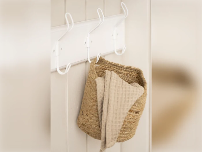 En flettet kurv og et beige tekstureret håndklæde hænger på hvide kroge på en cremefarvet væg.