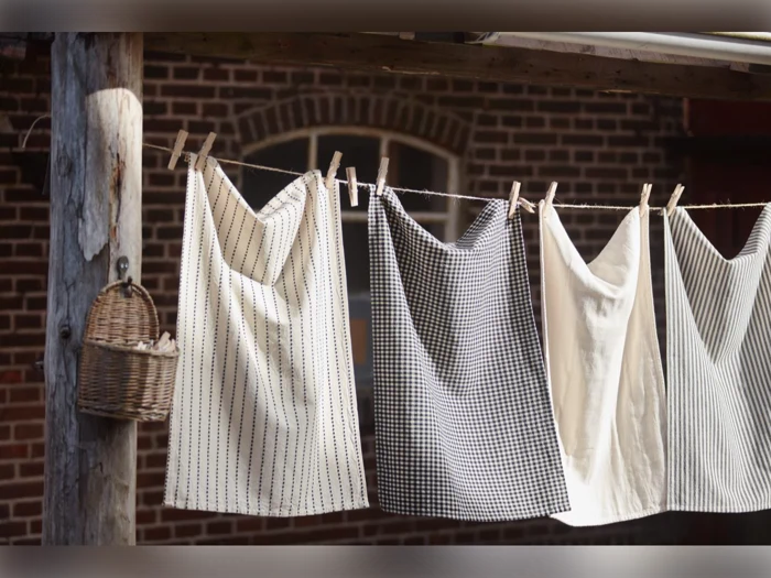 Fire håndklæder med forskellige mønstre hænger på en tørresnor med tøjklemmer udenfor, nær en træpæl og en lille flettet kurv. I baggrunden ses en murstensvæg.
