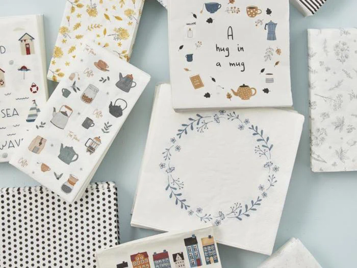 Et udvalg af mønstrede servietter med motiver af tekander, huse, blå blomsterkranse, polkaprikker, muslingeskaller og diskrete blomsterprint, arrangeret på en lys overflade.