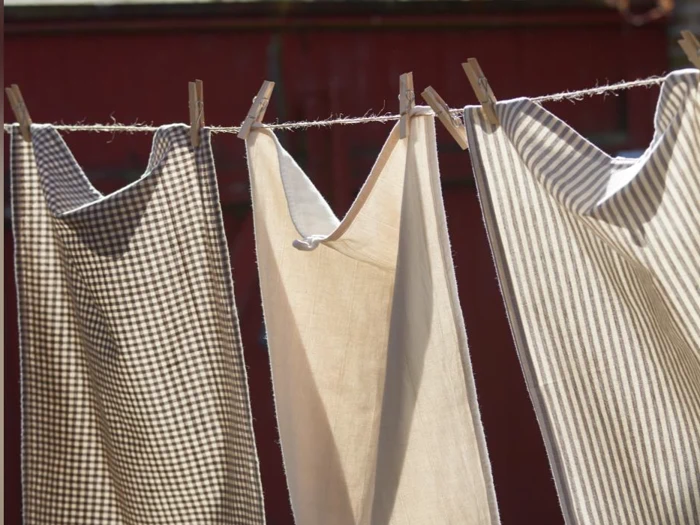 Tre stykker ternet og stribet stof hænger på en tørresnor med tøjklemmer af træ og tørrer i sollyset mod en sløret rød baggrund.