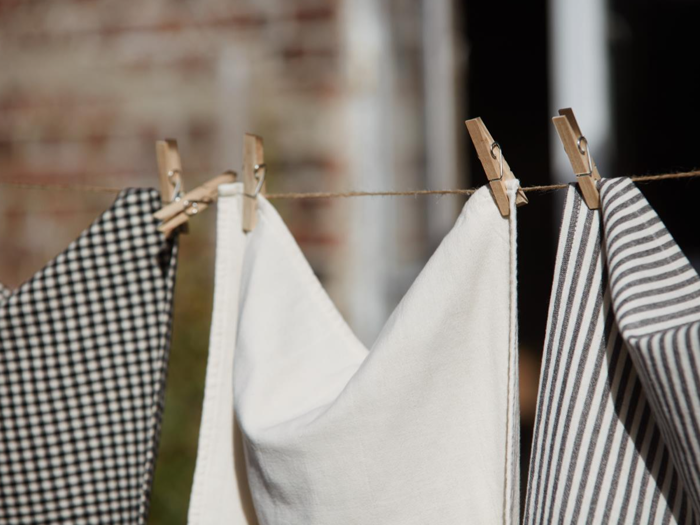 Tre stykker stof - et ternet, et almindeligt hvidt og et stribet - hænger på en tørresnor med tøjklemmer af træ udendørs.