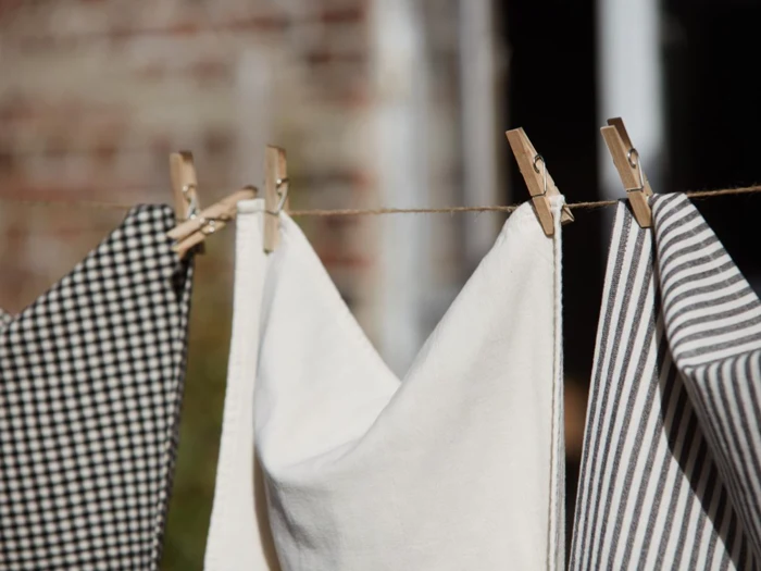 Tre stykker stof - et ternet, et almindeligt hvidt og et stribet - hænger på en tørresnor med tøjklemmer af træ udendørs.