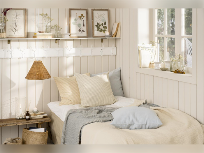 Hyggeligt soveværelse med en pænt redt seng, bløde pastelpuder, indrammede botaniske tryk, en hylde med dekoration og naturligt lys, der kommer ind gennem et vindue.