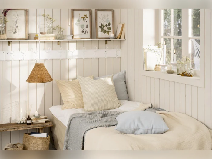 Hyggeligt soveværelse med en pænt redt seng, bløde pastelpuder, indrammede botaniske tryk, en hylde med dekoration og naturligt lys, der kommer ind gennem et vindue.