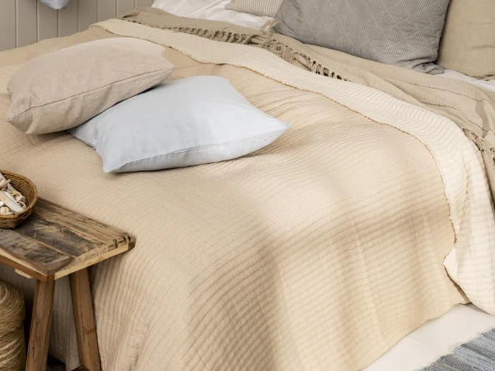 En pænt redt seng med beige og cremefarvede tæpper, en lyseblå pude og puder i neutrale farver ved siden af et rustikt sidebord i træ.