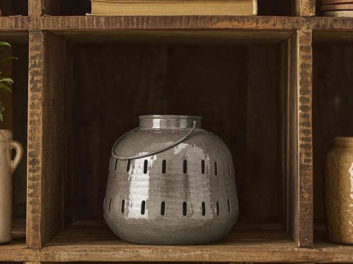En grå keramiklygte med lodrette udskæringer og et metalhåndtag står på en træhylde omgivet af rustikke dekorationsgenstande.