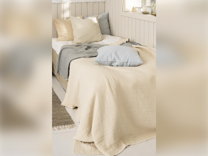 En hyggelig, pænt redt seng med cremefarvede, grå og beige puder og tæpper i et solbeskinnet soveværelse i neutrale farver.