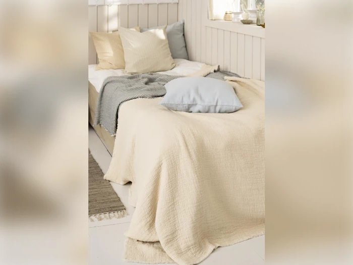 En hyggelig, pænt redt seng med cremefarvede, grå og beige puder og tæpper i et solbeskinnet soveværelse i neutrale farver.