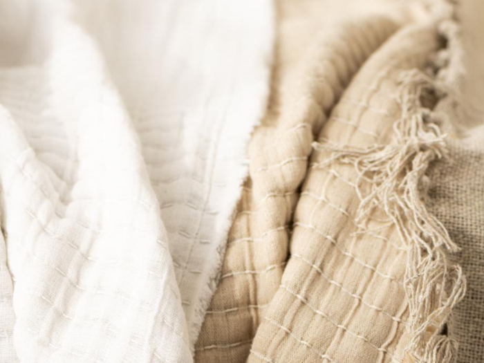 Blødt, struktureret stof i hvide og beige nuancer med synlige vævede mønstre og nogle flossede kanter, arrangeret i blide folder.
