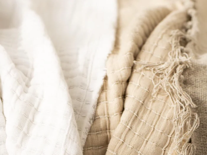 Blødt, struktureret stof i hvide og beige nuancer med synlige vævede mønstre og nogle flossede kanter, arrangeret i blide folder.