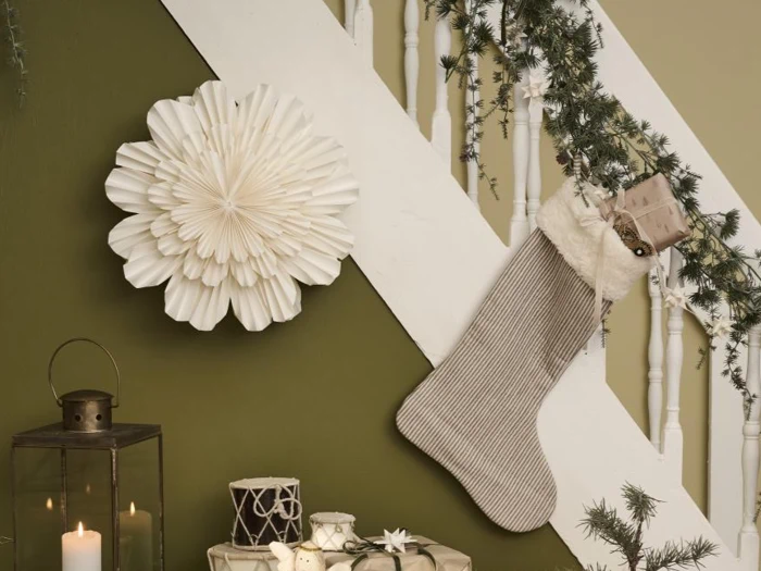 En trappe dekoreret med guirlande, en stor hvid papirblomst, en grå julestrømpe, stearinlys og indpakkede gaver på en grøn vægbaggrund.