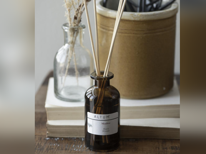 En brun glasflaske med sivpinde mærket ALTUM står på stablede bøger ved siden af en lille klar vase med tørrede blomster og en stor beige keramikbeholder.
