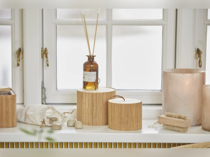 Bambusbeholdere, en brun glasflaske med sivpinde, sæbestykker, børster og en lysestage står i en hvid vindueskarm med blødt, naturligt lys.