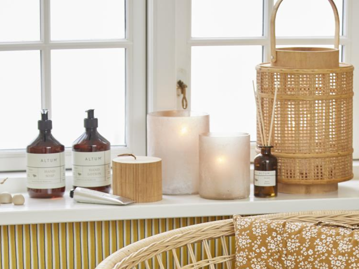 En hyggelig vindueskarm med håndsæbe, lotion, stearinlys, en flettet lanterne, små beholdere og en blomstret pude på en kurvestol.