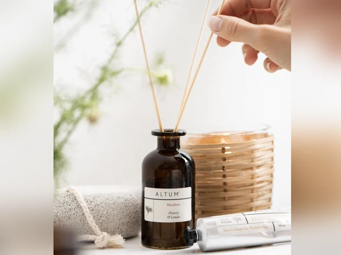 En hånd placerer siv i en brun ALTUM-diffusorflaske mærket Meadow, Honey & Lemon med et stearinlys, et rør og en sten i nærheden på en lys overflade.