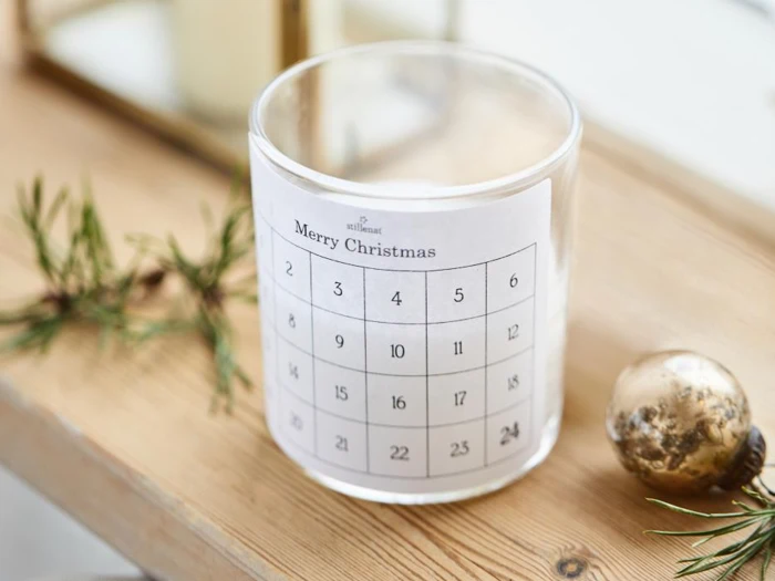 Et glas med en papirkalender med påskriften Glædelig jul viklet rundt om, placeret på et træbord med fyrrekviste og en guldkugle i nærheden.