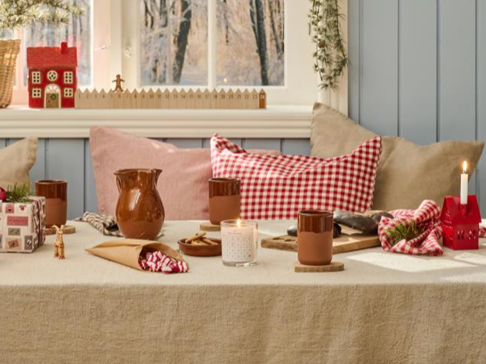 Et hyggeligt bord dækket med brune krus, en kande, ternede servietter, indpakkede gaver, et stearinlys og en lille husformet lysestage, med puder og et vindue i baggrunden.