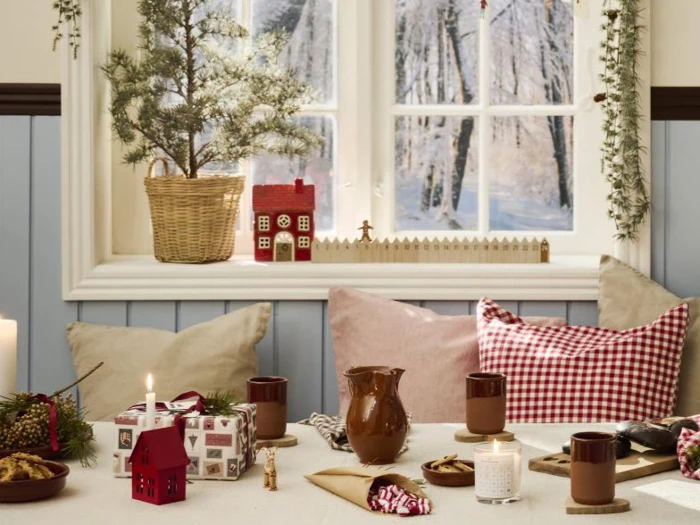 Et hyggeligt, festligt bord ved et vindue har stearinlys, krus, en kande, indpakkede gaver, småkager og julepynt, med et lille træ i en kurv og sneklædte træer udenfor.