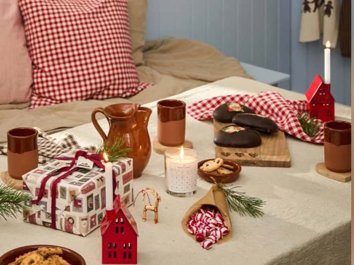 En hyggelig julescene med en seng, et festligt dækket bord med krus, småkager, stearinlys, indpakkede gaver, fyrretræsgrene og rødternede detaljer.