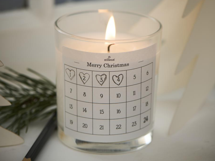 Et tændt stearinlys i en glasholder med en adventskalenderetiket med titlen Merry Christmas, der viser de nummererede dage 1 til 24. Fyrretræ og en blyant ligger i nærheden.
