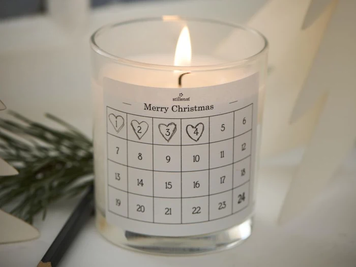 Et tændt stearinlys i en glasholder med en adventskalenderetiket med titlen Merry Christmas, der viser de nummererede dage 1 til 24. Fyrretræ og en blyant ligger i nærheden.