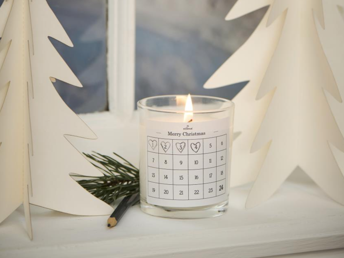 Et tændt stearinlys i en glasholder med design af en nedtællingskalender til jul, omgivet af hvide papirtræer og en fyrrekvist i en hvid vindueskarm.