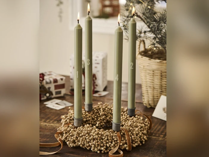 En adventskrans med fire grønne lys nummereret fra 1 til 4, omgivet af guldbær, står på et træbord med julepynt i baggrunden.