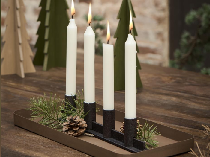 Fire tændte hvide stearinlys i sorte stager står på en brun bakke dekoreret med kogler og grønne planter, og træerne står i baggrunden på et træbord.