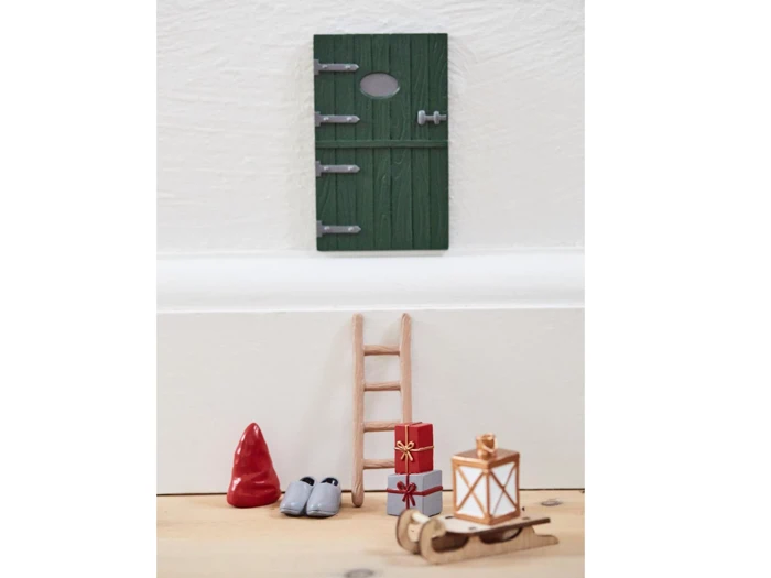 Grøn minidør på en hvid væg over en lille stige, rød hat, grå sko, indpakkede gaver, en lanterne og en lille træslæde på et trægulv.