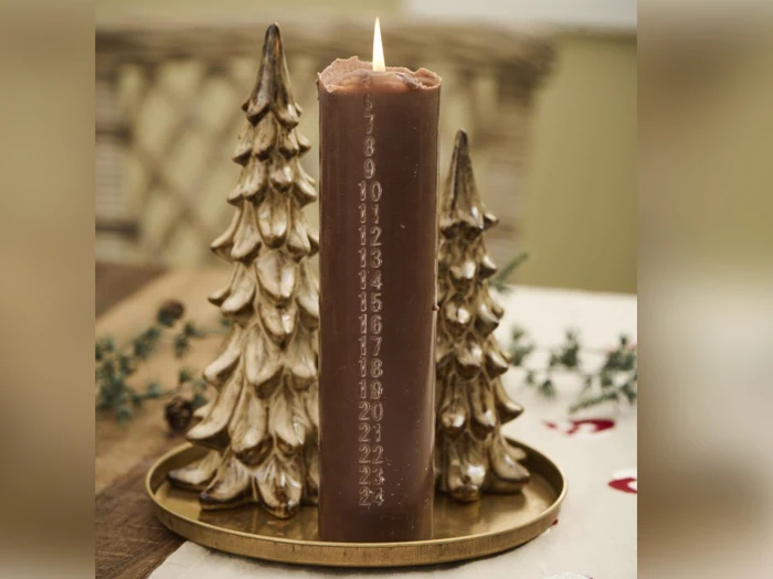 Et brunt adventslys med nummererede dage tændes og placeres på en guldbakke, flankeret af to dekorative guldjuletræsornamenter.
