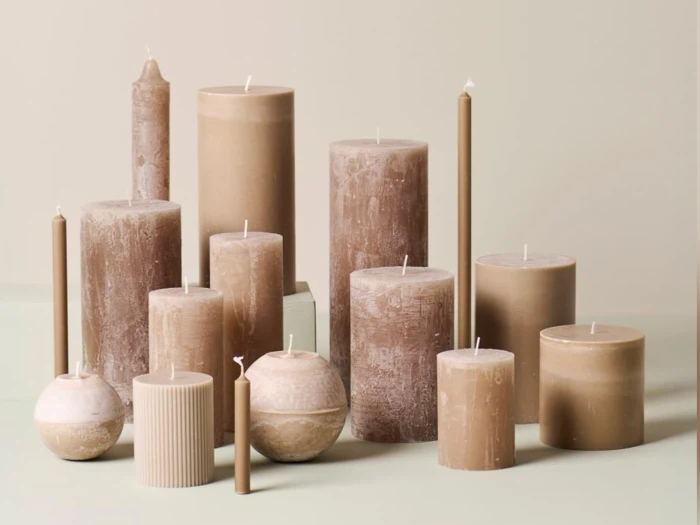 En samling af beige og brune stearinlys i forskellige former og størrelser er anbragt på en neutral overflade med en lys baggrund.