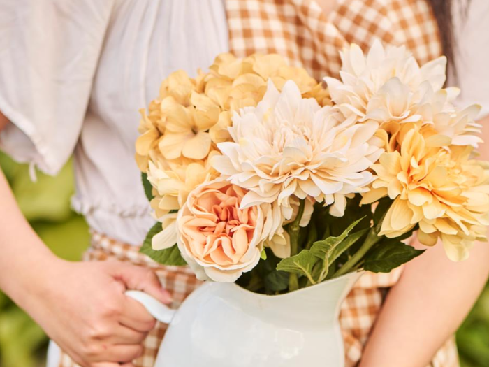 En person i gingham-kjole holder en hvid kande fyldt med cremefarvede og gule blomster, bl.a. hortensiaer og georginer.
