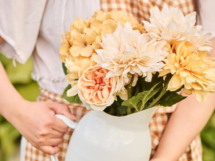 En person i gingham-kjole holder en hvid kande fyldt med cremefarvede og gule blomster med grønne blade.
