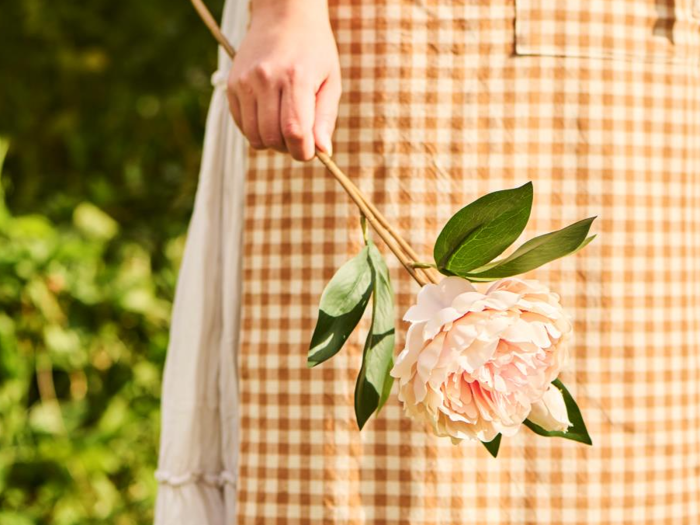 En person i gingham-kjole holder en lyserød pæonblomst med grønne blade mod en sløret udendørs baggrund.