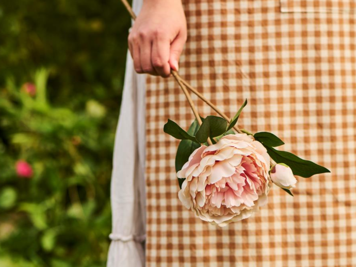 En person i en brun ternet kjole holder en lyserød pæonblomst i stilken, mens det grønne er sløret i baggrunden.