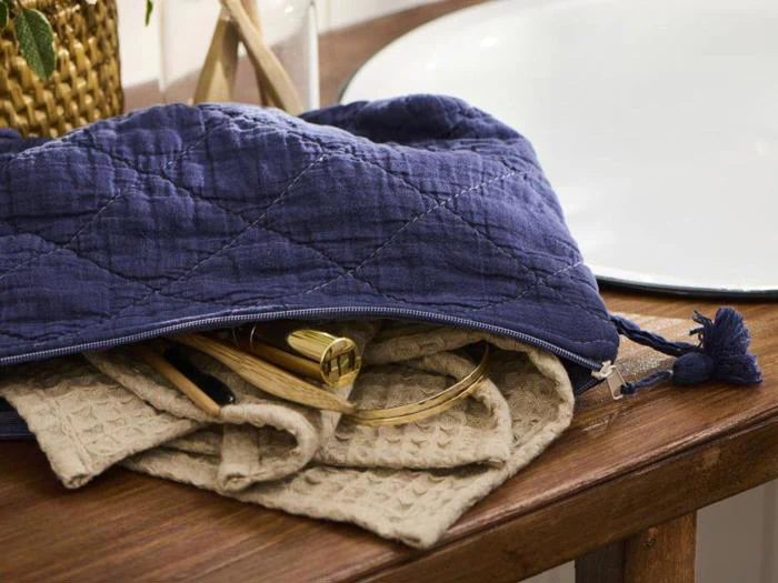 En blå quiltet lynlåspose på en træoverflade, delvist åben for at afsløre beige strukturerede klude og guldfarvede makeupbørster indeni.