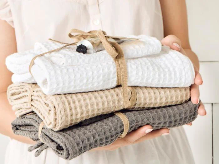 En person holder en stak foldede vaffelstrikkede håndklæder i hvid, beige og brun, bundet sammen med et solbrunt bånd og et lille rør på toppen.