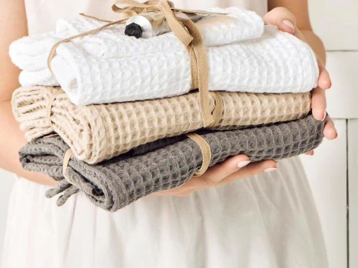 En person holder en stak foldede håndklæder med vaffelstruktur i hvid, beige og grå, bundet med et brunt bånd.