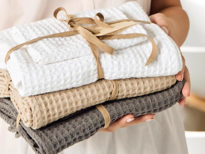 En person holder tre foldede håndklæder med vaffelstruktur i hvid, beige og grå, bundet sammen med et brunt bånd.