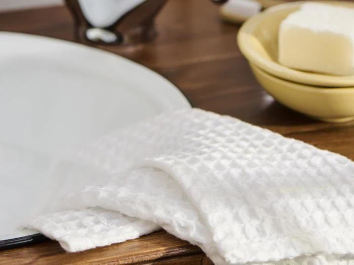 Et hvidt, struktureret håndklæde ligger ved siden af en hvid håndvask med en gul skål med et stykke sæbe på en træplade i baggrunden.